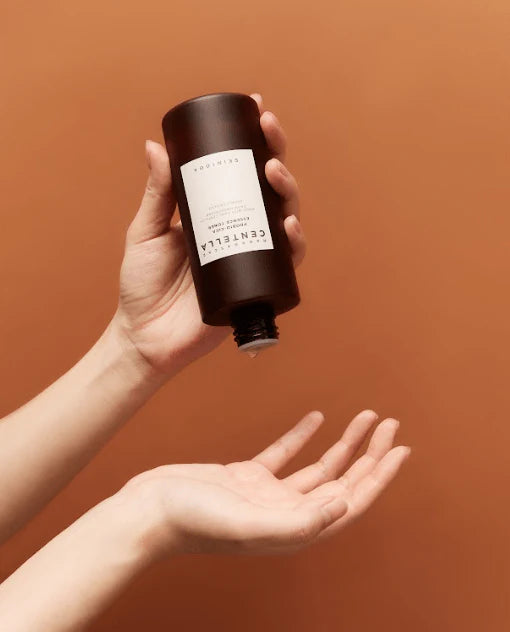 Hand holding a dark brown bottle labeled 'CENTELLE' against a warm brown background
